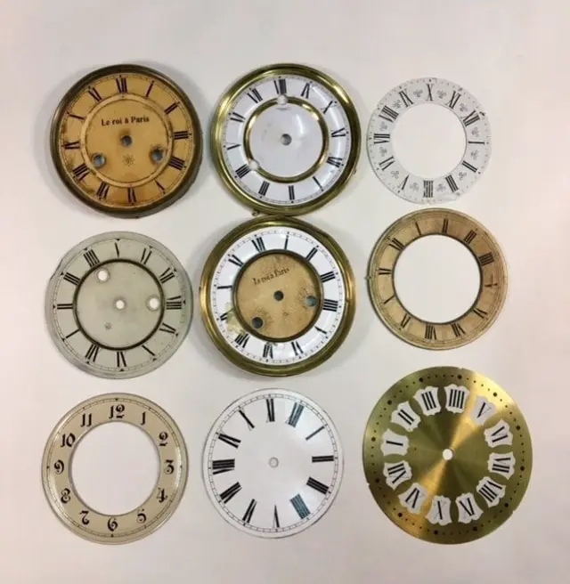 Zestaw vintage tarcz zegarowych w różnych stylach i wykończeniach, idealny do renowacji starych zegarów lub projektów DIY z antycznym charakterem. Na zdjęciu widoczne są tarcze z rzymskimi oraz arabskimi cyframi, wykonane z materiałów takich jak mosiądz, porcelana i metal, z widocznymi otworami montażowymi lub otwartym środkiem. Niektóre tarcze posiadają nadruki, np. Le roi à Paris, co dodaje im autentycznego, francuskiego stylu retro.