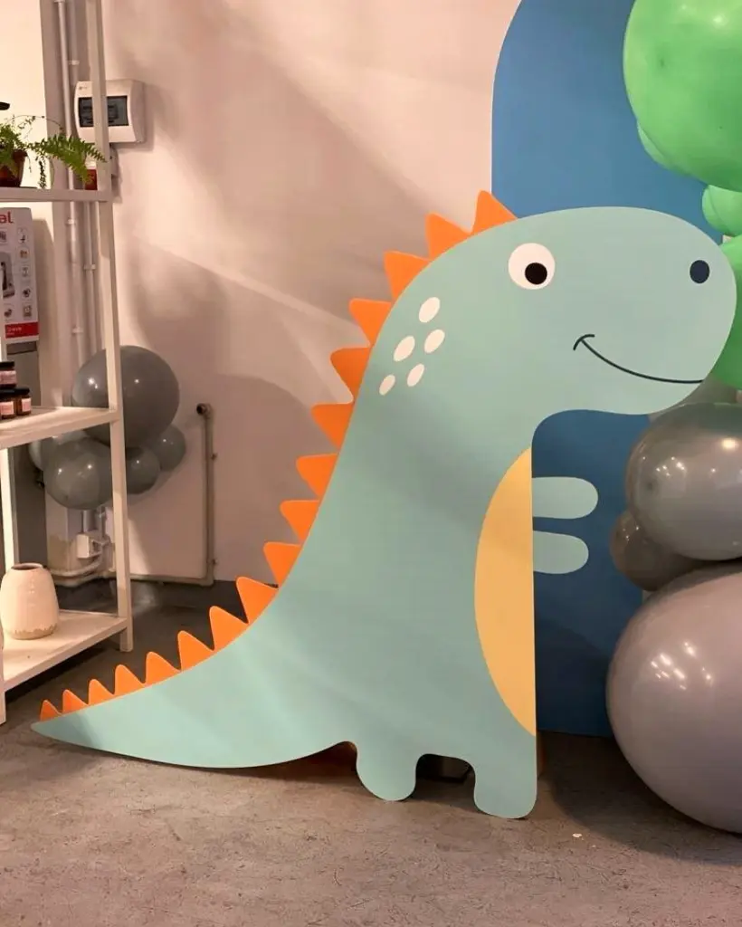 Dekoracja dziecięca przedstawiająca dużą, stojącą grafikę uśmiechniętego dinozaura w kolorze miętowym z pomarańczowymi kolcami na grzbiecie i żółtym brzuszkiem, ustawiona na podłodze wewnątrz pomieszczenia. W tle znajdują się balony w odcieniach szarości oraz niebieski panel dekoracyjny. Całość tworzy przyjazną, kolorową scenografię idealną na przyjęcie urodzinowe lub imprezę tematyczną dla dzieci