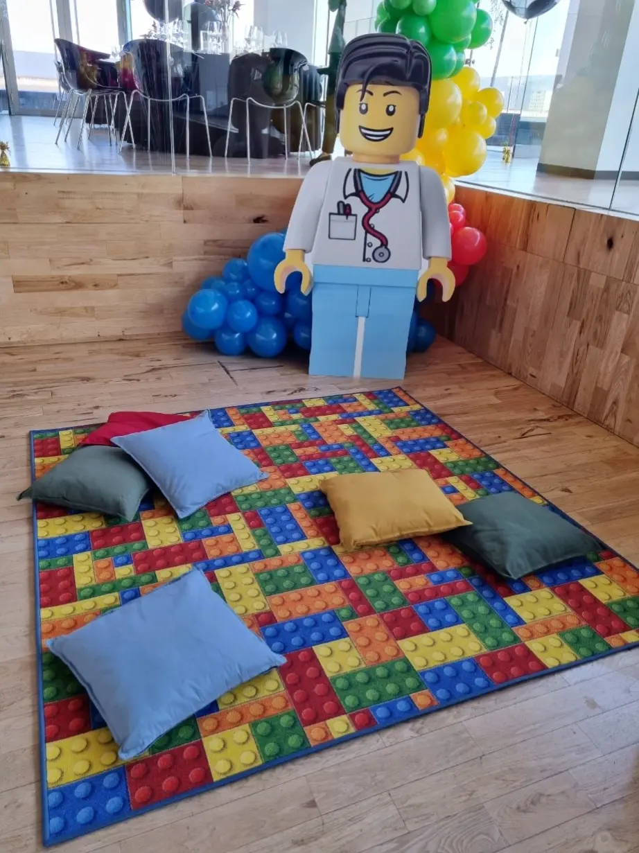 Strefa zabaw dla dzieci inspirowana klockami LEGO, z kolorowym dywanem przedstawiającym wzór z klocków w barwach czerwieni, niebieskiego, żółci, zieleni i pomarańczu. Na dywanie rozmieszczone są miękkie poduszki w różnych kolorach. W tle znajduje się duża tekturowa figura przedstawiająca postać LEGO lekarza z uśmiechem, stetoskopem i identyfikatorem. Dekorację dopełniają niebieskie i wielokolorowe balony ułożone przy ścianie oraz drewniana podłoga i szklana balustrada oddzielająca przestrzeń. Całość tworzy radosną i przyjazną przestrzeń idealną na urodziny lub event dla dzieci