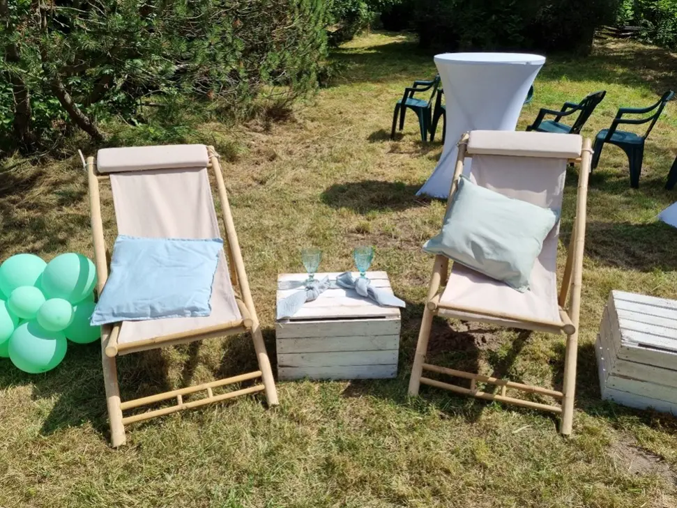 Strefa relaksu na świeżym powietrzu z dwoma leżakami z jasnobeżowym materiałem i pastelowymi poduszkami – niebieską i miętową. Pomiędzy nimi znajduje się biały, drewniany stolik skrzynkowy z dwiema szklankami i serwetkami. W tle widać wysoki stół koktajlowy z białym pokrowcem oraz zielone krzesła ogrodowe, a po lewej stronie jasne, zielone balony dekoracyjne ustawione na trawie. Całość zlokalizowana jest na trawiastym terenie w otoczeniu drzew