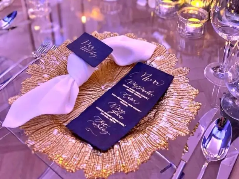 Eleganckie nakrycie stołu z przezroczystym talerzem i dekoracyjną złotą podkładką w kształcie płatków. Na talerzu znajduje się biały obrusowy pierścień, a obok niego menu w ciemnym kolorze z eleganckim, złotym nadrukiem w stylu calligraphy. Po bokach talerza ułożone są srebrne sztućce, a w tle widać kieliszki do wina oraz małe, zapalone świeczki, tworząc nastrojową, romantyczną atmosferę na uroczystość