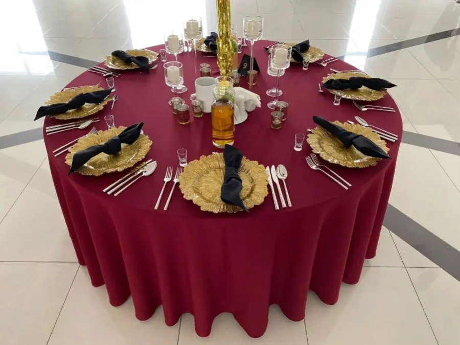Stół przykryty jest eleganckim bordowym obrusem. Na talerzach w kształcie złotych liści znajdują się czarne serwetki, starannie złożone w kokardy. Wokół stołu rozmieszczono srebrne sztućce oraz kieliszki, a także małe szklane świeczniki, które dodają subtelnego blasku. Centralnym punktem dekoracyjnym jest wysoka złota świeca oraz butelka z napojem, otoczona przez mniejsze szklane kieliszki. Całość emanuje prostą, ale wyrafinowaną elegancją, tworząc atmosferę odpowiednią na wyjątkową okazję