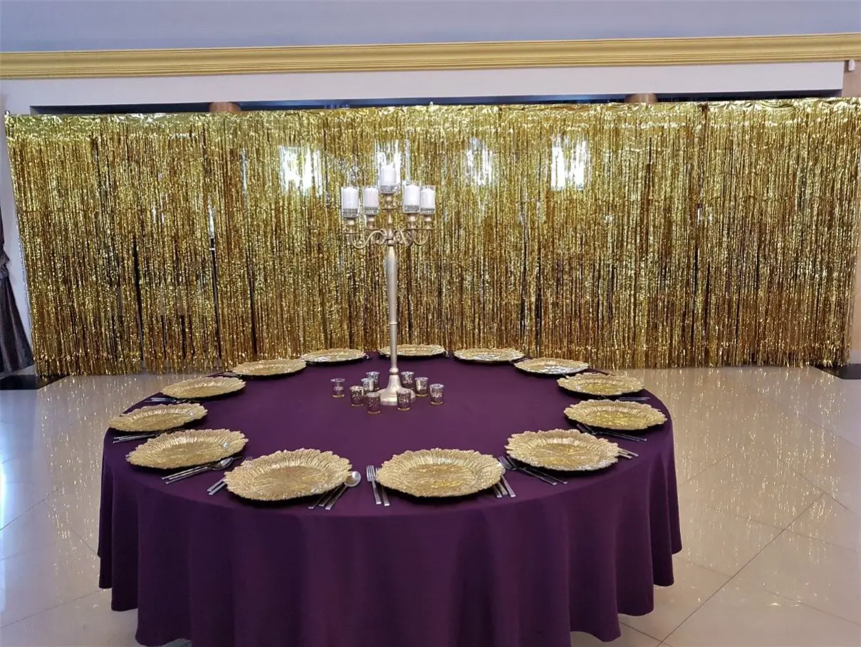 Stół przykryty jest głębokim fioletowym obrusem, a na nim znajdują się talerze w kształcie złotych liści, które stanowią elegancki akcent. Wokół talerzy rozstawione są srebrne sztućce, a na stole ustawiono świeczniki i małe szklane świeczki, które wprowadzają ciepłe światło. W tle wisi błyszcząca złota zasłona, która dodaje blasku i glamour całej aranżacji. W centrum stołu znajduje się wysoki świecznik, tworząc elegancką i luksusową atmosferę, idealną na specjalne okazje
