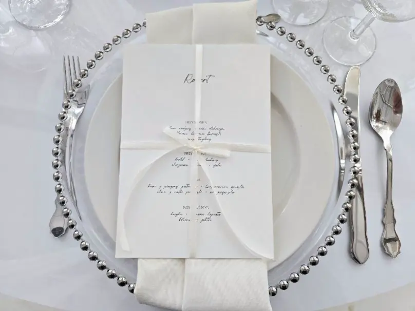 Eleganckie nakrycie stołu z przezroczystym talerzem ozdobionym srebrnym koralikowym obrusem. Na talerzu znajduje się starannie ułożone menu, zawiązane białą wstążką, oraz biały serwetka. Po bokach talerza umieszczone są srebrne sztućce – widelec, nóż i łyżka. W tle widoczne są kieliszki do wina, tworząc subtelną, elegancką atmosferę na wyjątkową okazję, taką jak kolacja lub wesele