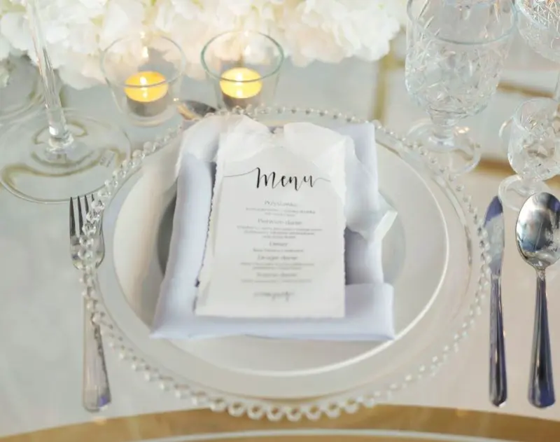 Eleganckie nakrycie stołu z przezroczystym talerzem ozdobionym srebrnym koralikowym brzegiem. Na talerzu znajduje się jasnoniebieska serwetka z zawiniętym w nią menu, nadrukowanym eleganckim, ręcznym pismem. W tle widoczne są kieliszki do wina, a obok talerza zapalone małe świeczki w szklanych pojemnikach, tworzące romantyczną atmosferę. Całość prezentuje się delikatnie i stylowo, idealna na uroczystość w eleganckim wydaniu