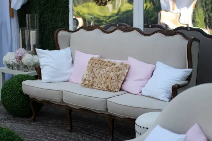 Stylowa, beżowa sofa z ciemnym, drewnianym wykończeniem ustawiona na brukowanej powierzchni w eleganckim ogrodowym otoczeniu. Na sofie ułożono dekoracyjne poduszki w odcieniach bieli, pudrowego różu i beżu, w tym jedną z trójwymiarowymi kwiatowymi aplikacjami. W tle widoczne są zielone ozdobne kule z bukszpanu, świece w szklanych cylindrach oraz biały stolik z bukietami jasnych kwiatów