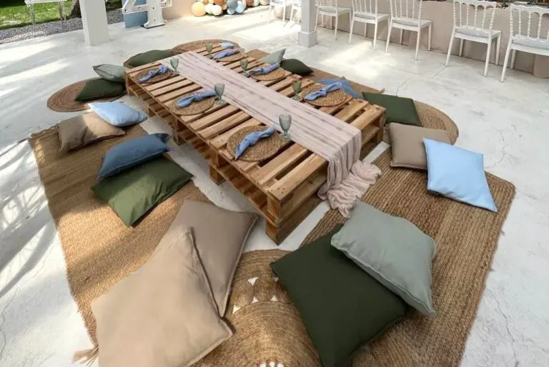 Niskie, rustykalne stoły wykonane z drewnianych palet ustawione na naturalnych, plecionych dywanach w stylu boho. Wokół nich rozłożone są duże poduszki w odcieniach zieleni, beżu, błękitu i szarości, służące jako siedziska. Na stole znajdują się plecione podkładki, szklane kielichy i starannie ułożone lniane serwetki w odcieniach niebieskiego. W tle białe krzesła i pastelowe dekoracje, sugerujące przyjęcie lub uroczystość w plenerze