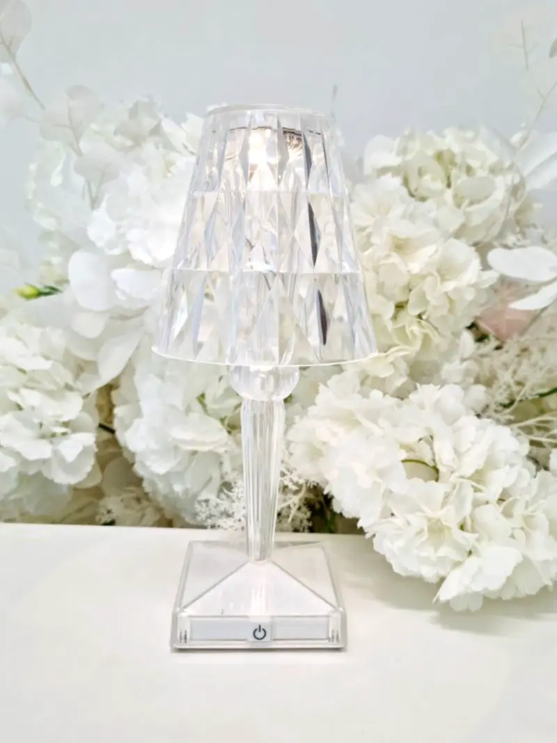 Elegancka, przezroczysta lampka stołowa o diamentowym szlifie, wykonana z tworzywa przypominającego kryształ. Lampka stoi na białej powierzchni, a w tle widoczna jest dekoracja z dużych, białych hortensji i delikatnych liści, tworząca romantyczną i subtelną atmosferę. Na podstawie lampki znajduje się mały przycisk z symbolem włącznika