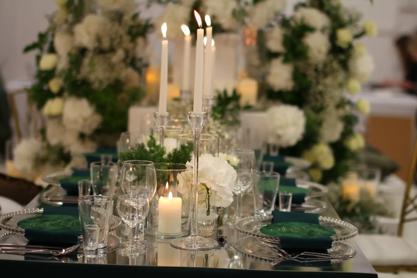 Elegancko nakryty stół weselny z lustrem jako blatem, ozdobiony wysokimi, smukłymi świecznikami ze świecami w kolorze białym oraz szklanymi naczyniami z palącymi się świecami. Nakrycia stołowe zawierają przezroczyste talerze z dekoracyjnym brzegiem oraz ciemnozielone serwetki ułożone na środku. W tle znajdują się bujne kompozycje z białych hortensji i zielonych liści, tworzące romantyczną, kwiatową ramę wokół stołu