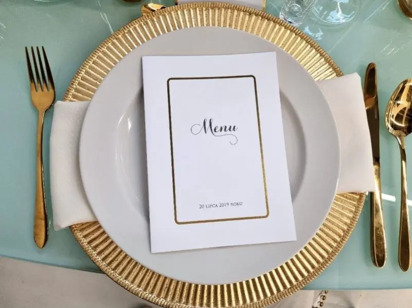 Elegancko nakryty stół z białym talerzem na złotej podkładce. Na talerzu znajduje się menu z napisem „Menu” w ozdobnym, kaligraficznym fontem, oprawione złotą ramką. Pod kartą leży biała serwetka, złożona w prosty sposób, a obok talerza znajdują się złote sztućce: widelec po lewej stronie oraz nóż i łyżka po prawej. W tle widać delikatnie błękitny obrus, nadający subtelnego charakteru całemu nakryciu. Na stole widoczny jest także kieliszek do wina, który dopełnia elegancką aranżację