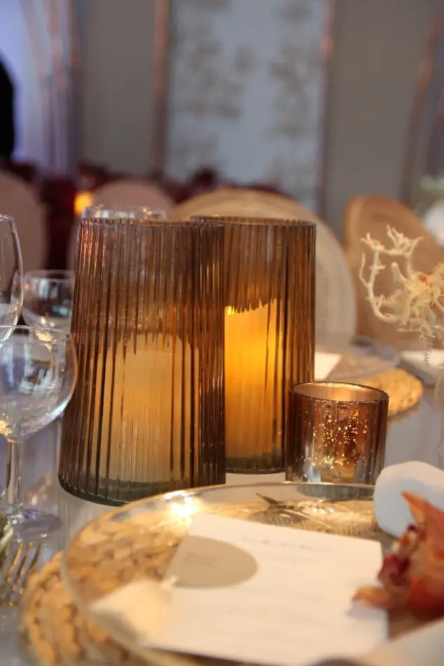 Zbliżenie na elegancką dekorację stołu złożoną z dwóch karbowanych szklanych lampionów w kolorze bursztynowym z zapalonymi świecami wewnątrz, obok których znajduje się mniejszy, metaliczny świecznik tealight. Całość umieszczona jest na złotym, plecionym podtalerzu z białym menu weselnym, tworząc ciepłą, nastrojową atmosferę idealną na romantyczne przyjęcie lub kolację w stylu glamour