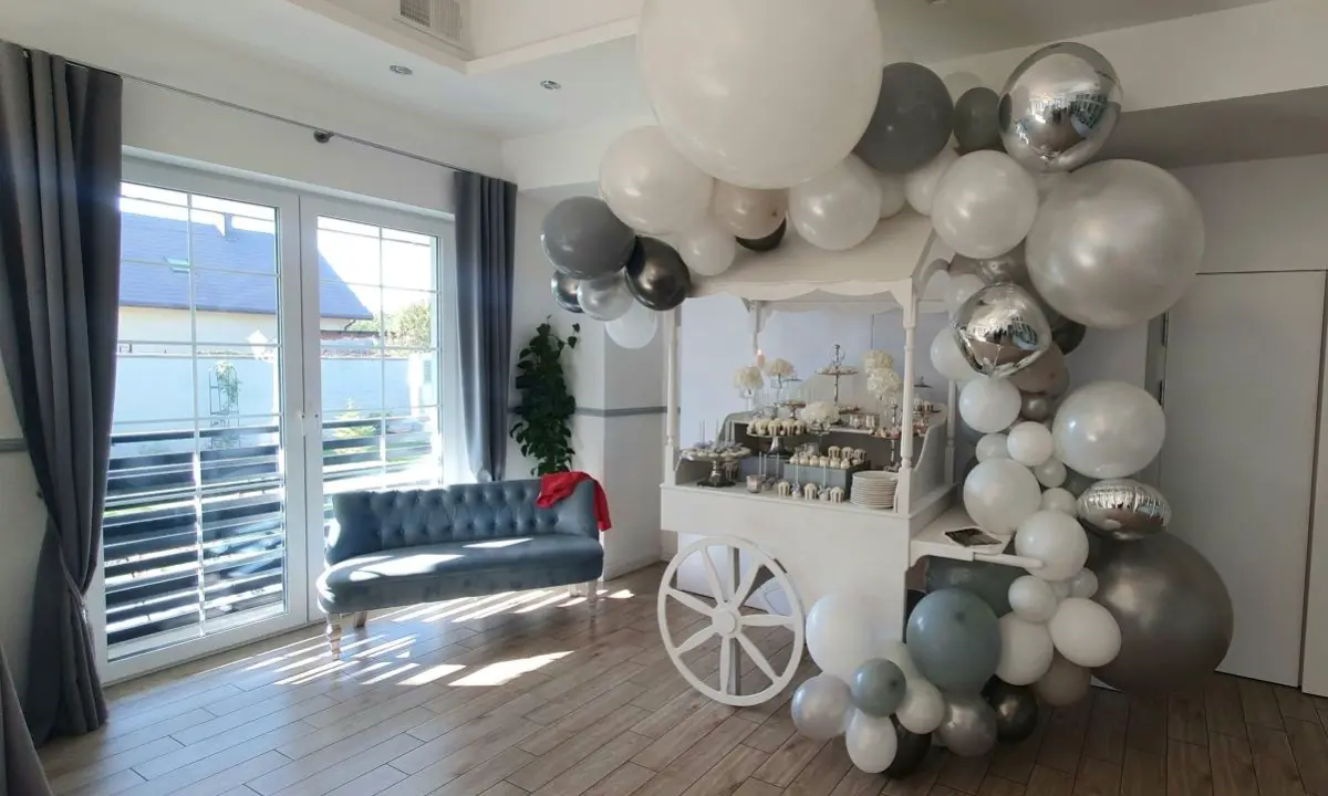 Nowoczesna aranżacja candy baru w stylu glamour z dominacją srebra, bieli i szarości. Centralnym elementem jest biały wózek w stylu retro, udekorowany efektowną girlandą z balonów w różnych odcieniach metalicznej szarości i bieli, w tym balonami chromowanymi. Na wózku ustawione są eleganckie desery, srebrne tace, białe kwiaty i ozdobne patery. Całość znajduje się w jasnym wnętrzu z drewnianą podłogą i dużymi oknami, które doświetlają przestrzeń naturalnym światłem. Obok widoczna jest stylowa sofa z pikowaniem i dekoracyjny czerwony element dodający kontrastu
