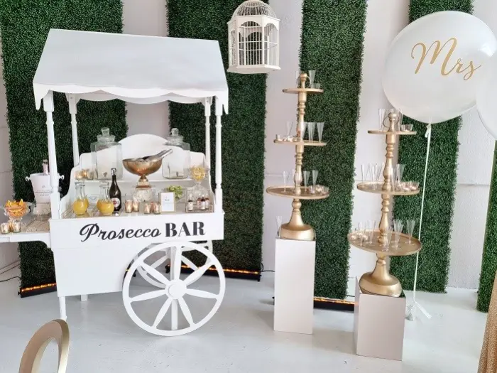 Elegancki prosecco bar w stylu weselnym, z białym wózkiem dekoracyjnym ozdobionym napisem „Prosecco BAR”. Na wózku ustawione są kieliszki, butelki prosecco, karafki z sokami oraz miska z lodem. Obok znajdują się dwie wysokie złote etażerki na białych cokołach, na których rozstawione są kieliszki do prosecco. W tle zielona ściana z paneli z bukszpanu, zawieszona dekoracyjna klatka i biały balon z napisem „Mrs” w złotym kolorze. Stylizacja idealna na wesele, wieczór panieński lub eleganckie garden party