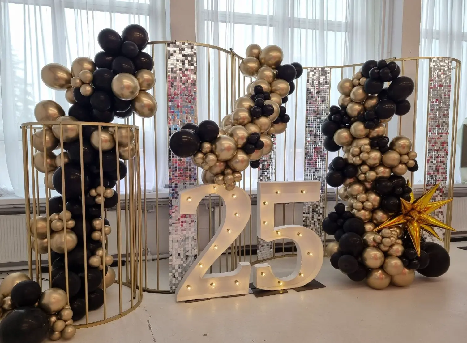 Efektowna dekoracja jubileuszowa na 25-lecie, utrzymana w kolorystyce czerni, złota i bieli. Tło stanowią złote metalowe stelaże z lustrzanymi, błyszczącymi panelami oraz okazałe girlandy balonowe z czarnych i złotych balonów w różnych rozmiarach. Centralnym punktem jest duży, podświetlany napis „25” w białym kolorze. Po prawej stronie dekorację uzupełnia złota, błyszcząca gwiazda 3D. Całość tworzy nowoczesną, glamour oprawę idealną na elegancki jubileusz, rocznicę lub sylwestrową imprezę