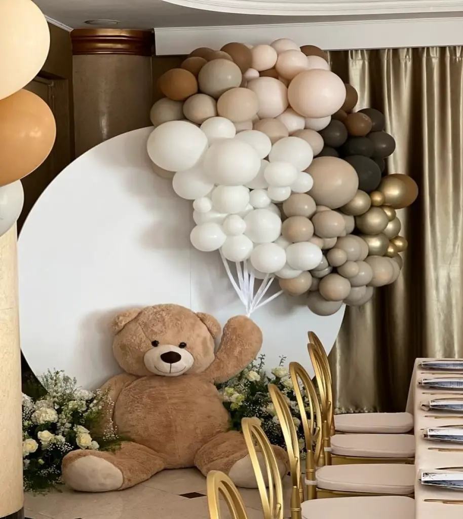 Dekoracja na przyjęcie z dużym pluszowym misiem trzymającym biały bukiet balonów w odcieniach beżu, brązu, bieli i złota, ustawiona na tle białego, okrągłego panelu. Całość uzupełniona złotymi krzesłami, białym obrusem i kompozycjami kwiatowymi z białych róż. Uroczy i elegancki wystrój idealny na baby shower, urodziny dziecka lub chrzest w stylu neutralnym i glamour