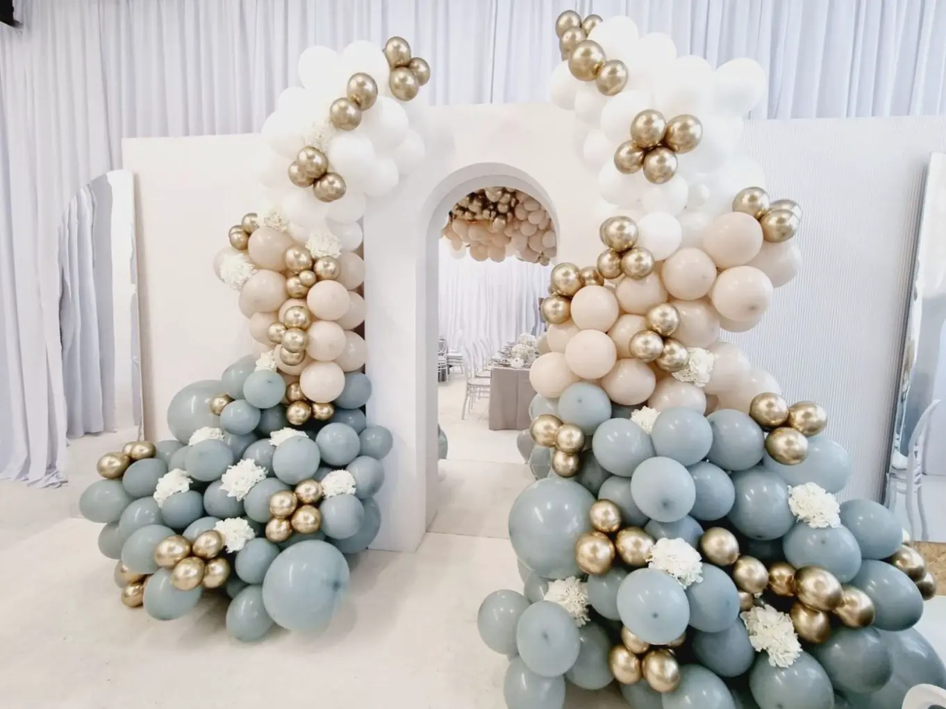 Elegancka dekoracja z balonów przy białym łukowym przejściu, składająca się z pastelowych balonów w kolorach szarym, beżowym i białym oraz złotych balonów chromowanych, tworzących efektowne akcenty. Wplecione białe kwiaty dodają dekoracji subtelności. Styl glamour, pastelowa kolorystyka i złote dodatki sprawiają, że aranżacja idealnie nadaje się na baby shower, roczek, chrzest lub eleganckie urodziny. Tło z białych zasłon i prześwitująca sala z dekoracją w tle dodają głębi kompozycji