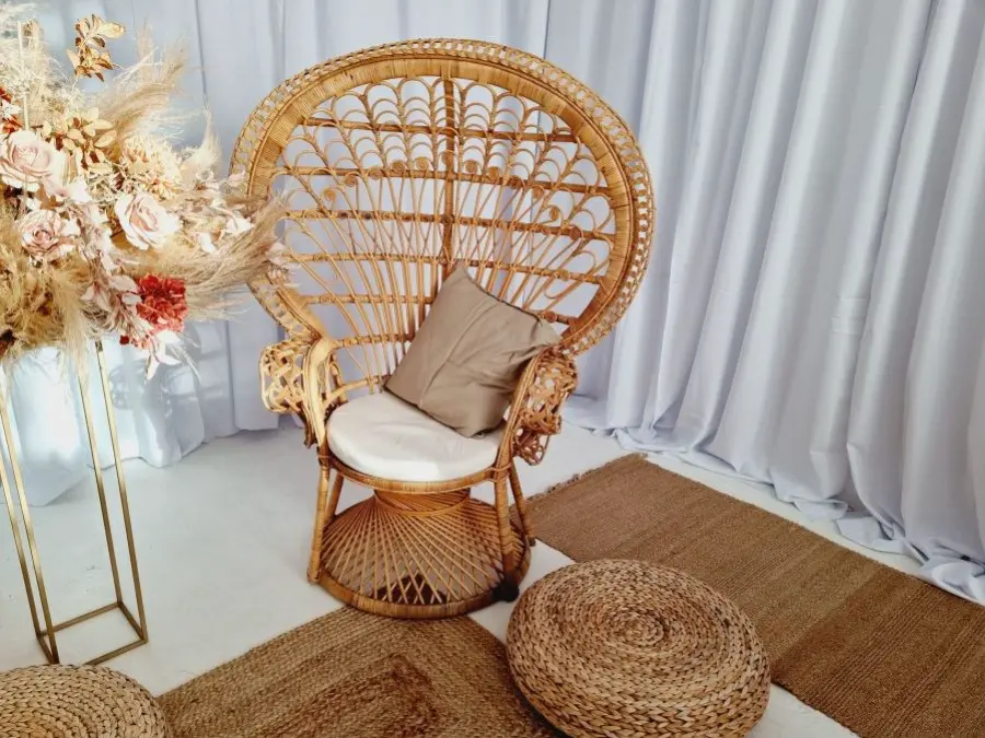 Stylowa aranżacja boho z dużym rattanowym fotelem typu „peacock chair” ozdobionym beżową poduszką, ustawionym na naturalnych jutowych dywanach. Przed fotelem dwa plecione pufy, a po lewej stronie złoty stojak z efektowną kompozycją z suszonych traw, liści palmowych i kwiatów w odcieniach beżu, różu i czerwieni. Tło stanowią jasnoszare, falujące zasłony, tworzące eleganckie, eteryczne tło. Idealna dekoracja na wesele, sesję zdjęciową lub event w stylu boho i rustykalnym