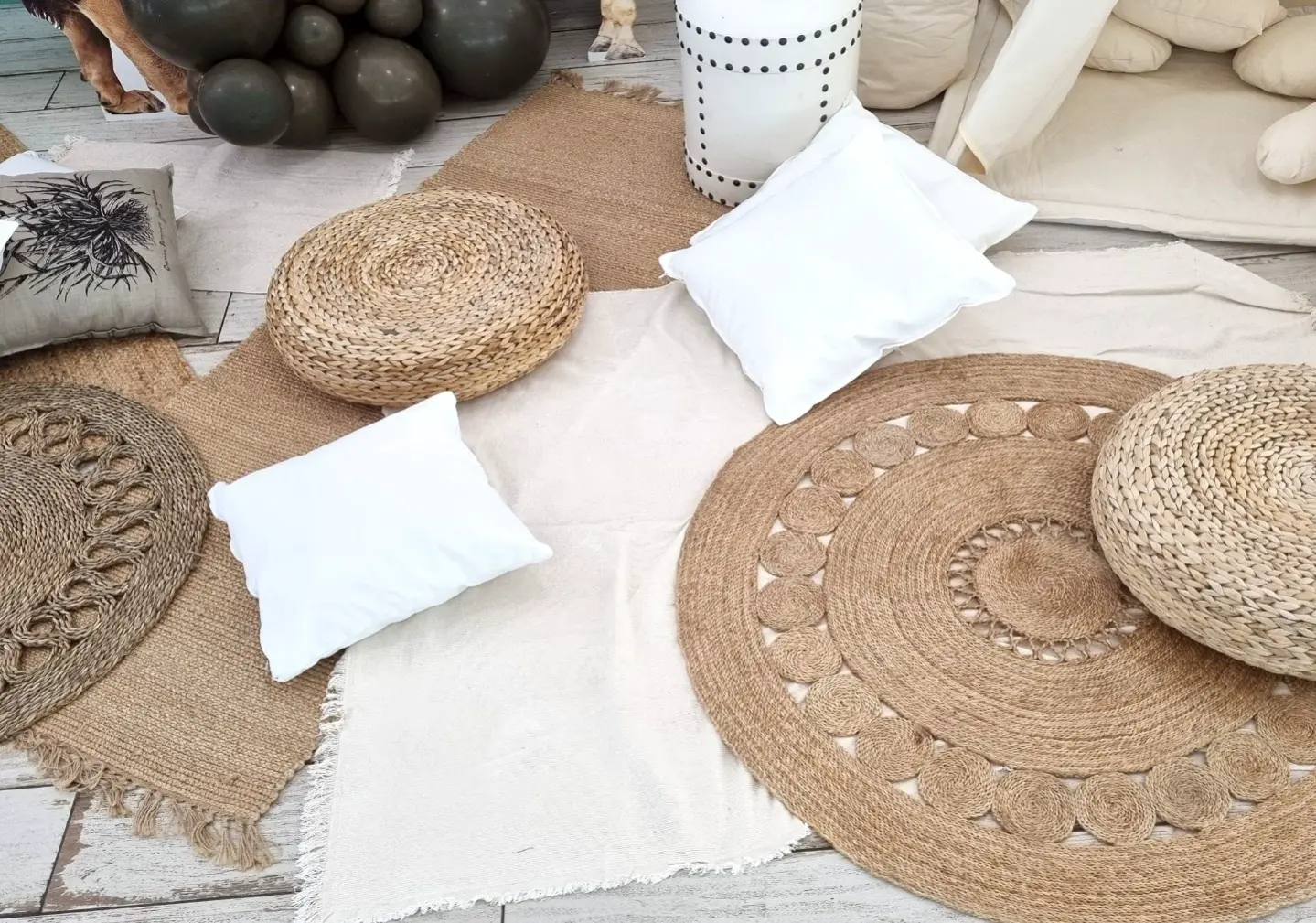 Przytulna strefa wypoczynkowa w stylu boho zaaranżowana na podłodze z jasnych, przecieranych paneli. Na powierzchni ułożono różnorodne jutowe i bawełniane dywany w naturalnych odcieniach beżu, brązu i bieli. Na nich rozmieszczono dekoracyjne plecione pufy z trawy morskiej oraz białe i szare poduszki, w tym jedna z czarnym nadrukiem roślinnym. Całość dopełniają ciemne balony w tle i jasne tkaniny, tworząc przyjazną atmosferę idealną na relaks, piknik, sesję zdjęciową lub przyjęcie w stylu boho