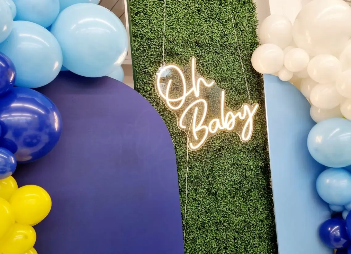 Dekoracja na baby shower w chłopięcej kolorystyce z napisem „Oh Baby” w formie jasnego neonu zawieszonym na zielonej ściance z liści. Po bokach znajdują się kolorowe panele w odcieniach niebieskiego oraz girlandy z balonów w kolorach niebieskim, białym i żółtym. Aranżacja tworzy radosną, nowoczesną scenerię idealną na przyjęcie z okazji narodzin dziecka, sesję zdjęciową lub baby shower w stylu playful i vibrant