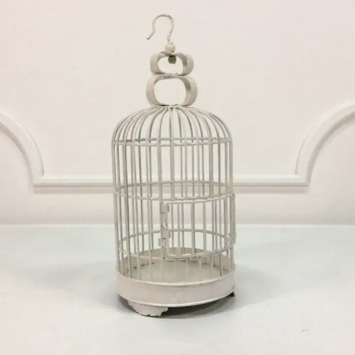 Okrągła dekoracyjna klatka w kolorze postarzanego bieli, wykonana z metalu i wyposażona w ozdobny uchwyt z haczykiem do zawieszenia. Posiada delikatne przetarcia w stylu shabby chic oraz zaokrąglone nóżki, nadające jej elegancki, rustykalny charakter. Idealna jako element dekoracyjny na wesele, świecznik, pojemnik na kartki z życzeniami lub romantyczna ozdoba do wnętrz