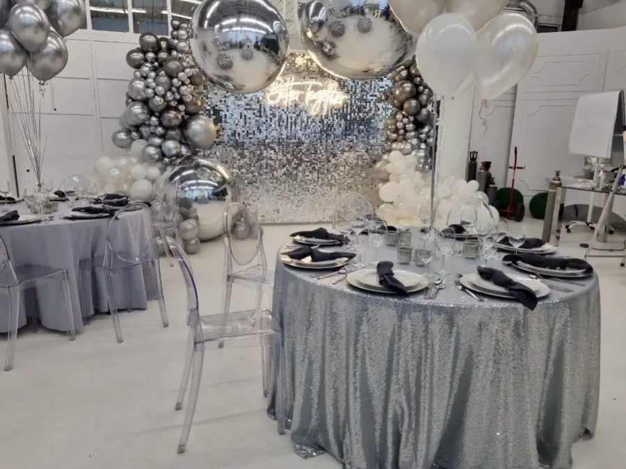 Nowoczesna aranżacja stołu na eleganckie przyjęcie, z błyszczącym, srebrnym obrusem i eleganckimi, przezroczystymi krzesłami. Na stole znajdują się białe talerze z ciemnymi serwetkami, kieliszki do wina oraz srebrne sztućce, tworząc minimalistyczną, ale wytworną dekorację. W tle widać efektowną dekorację balonową w odcieniach srebra, bieli i szarości, tworzącą nowoczesną atmosferę. Za nią wyróżnia się lustrzana ściana z migoczącymi płytkami oraz neonowym napisem Let’s Celebrate. Całość emanuje elegancją, idealną na jubileusze, urodziny lub inne uroczystości