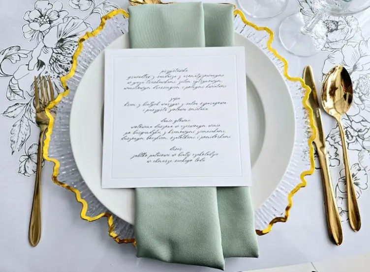 Elegancko nakryty stół z przezroczystym talerzem ozdobionym złotym rantem. Na talerzu znajduje się jasnozielona serwetka złożona w elegancki sposób oraz menu z napisanym ręcznie tekstem w stylu calligraphy. Po bokach talerza leżą złote sztućce – widelec, nóż oraz łyżka. Tło zdobi subtelny, biały obrus z delikatnym kwiatowym wzorem, tworzący klasyczną i wyrafinowaną atmosferę